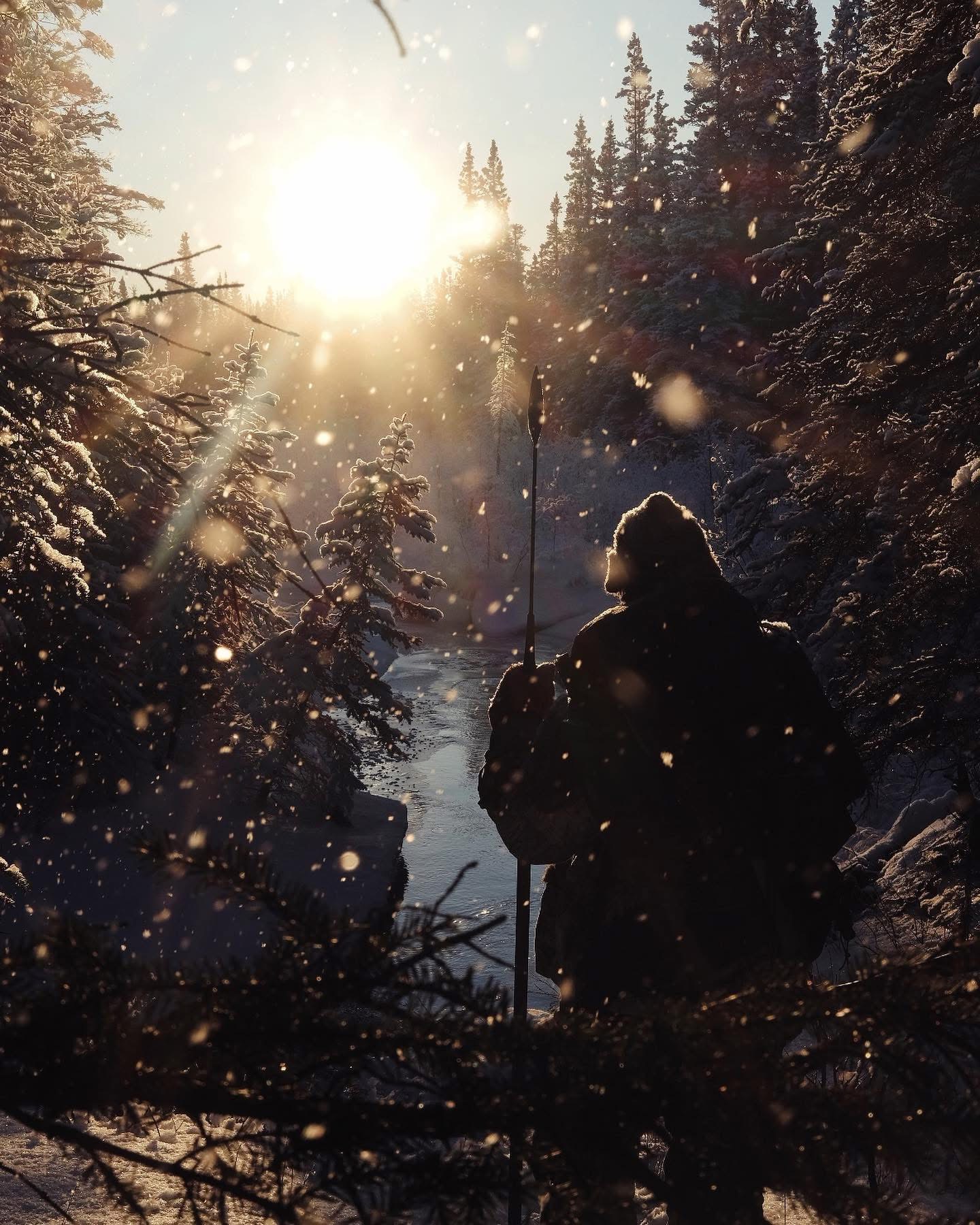 A person walks through a snowy Alaskan forest carrying a long tool, silhouetted against low sunlight and drifting snow.