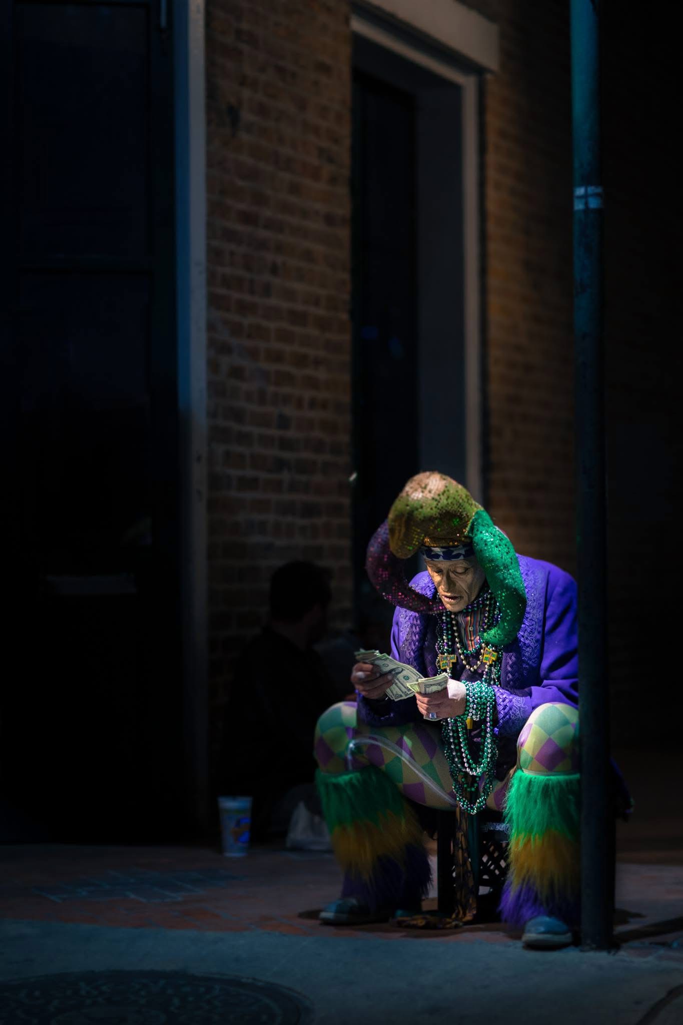 A seated person in a colorful jester costume counts money in a pool of light against a shadowed brick facade.